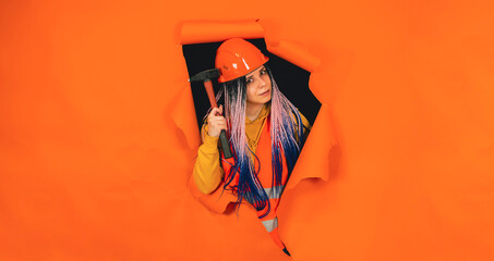 A female construction worker thinks and scratches her helmet with a hammer, trying to remember something. A woman in an orange protective helmet and vest
