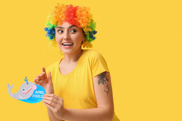 Funny young woman in colorful wig and with paper fish on yellow background. April fools' day...