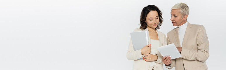 Obraz premium Businesswoman holding digital tablet near colleague with paper folder isolated on grey, banner.