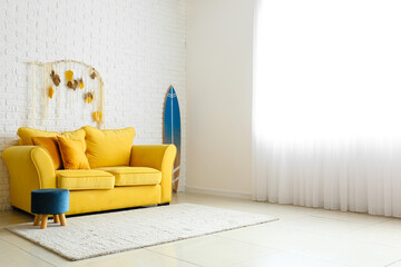 Comfortable sofa, pouf and surfboard near white brick wall in light room interior