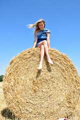Naklejka na ściany i meble Young attractive blonde woman sits barefoot on a round haystack and smiles