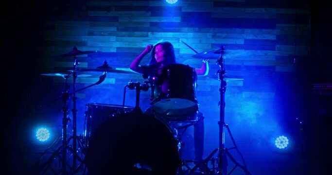 Drummer girl energetically playing the drums in blue, red and green twinkling strobe lights. Drummer girl concert concept. Camera moves away from the girl.