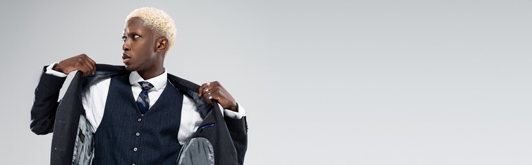 blonde african american man looking away and adjusting blazer isolated on grey, banner
