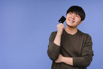 Pensive thoughtful asian korean man boy student freelancer holding credit card, dreaming about buying online shopping presents, cashback e-commerce, loan, scholarship isolated in blue