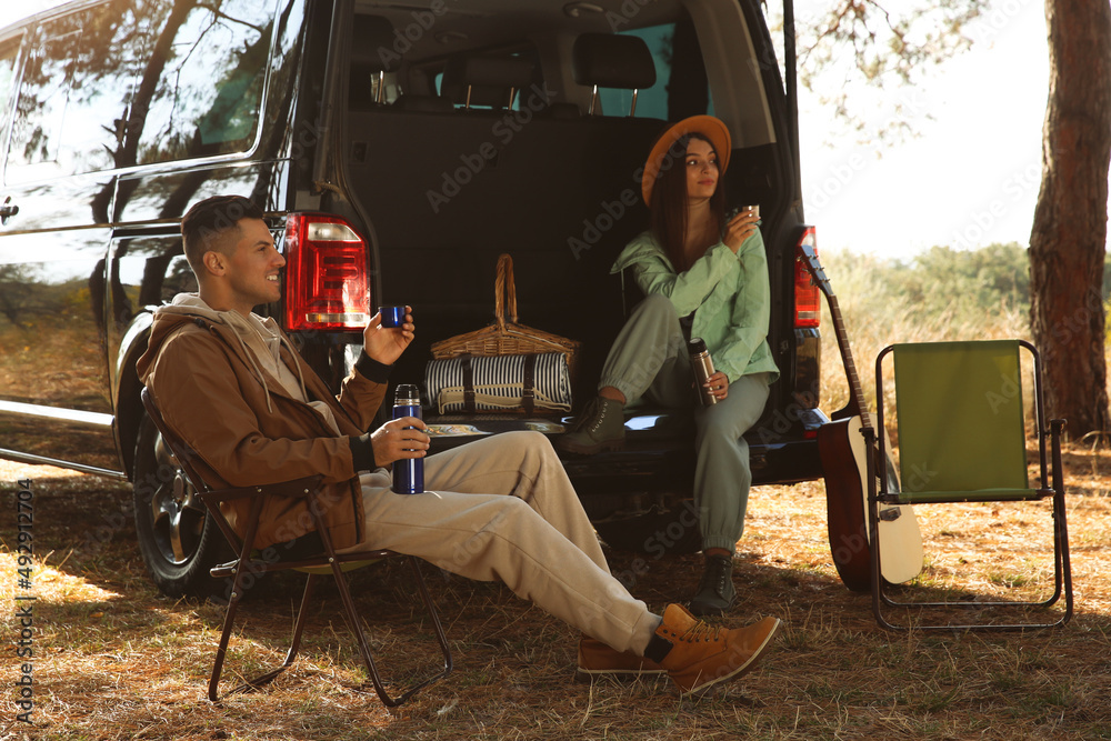 Wall mural Couple resting near car and enjoying hot drink at camping site