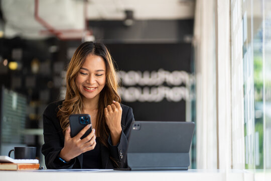 Smiling Asian businesswoman using phone in office. Small business entrepreneur looking at her mobile phone and smiling.