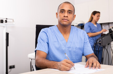 Obraz premium Positive Hispanic doctor cosmetologist in blue overall filling out medical form with female assistant preparing equipment for procedures on background..