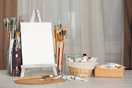 Many brushes, easel with blank canvas and paints on white wooden table indoors