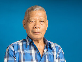 An elderly Asian man in a blue shirt looking at the camera with a smile while standing on a blue background