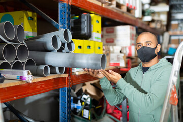 Craftsman in protective mask selects plastic plumbing pipes in a hardware store