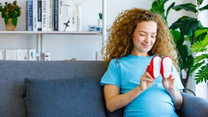 Caucasian happy pretty long curly hair healthy pregnancy mother in casual outfit sitting smiling on cozy sofa holding looking at small baby red shoes pair in living room expecting for newborn child