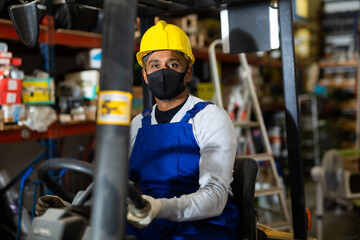 Portrait of confident hispanic worker in protective face mask driving forklift in building materials store. Concept of work in pandemic