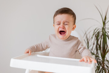 sad upset baby is sitting in a white high chair for feeding. little boy is crying