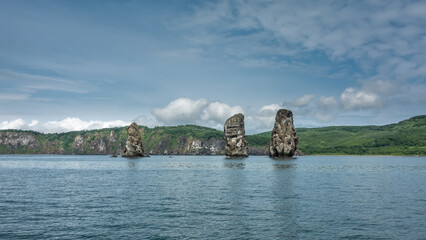 Fototapeta na wymiar High picturesque cliffs with steep slopes rise above the Pacific Ocean. The green hilly coast of Kamchatka against the background of blue sky and clouds. Rocks Three Brothers