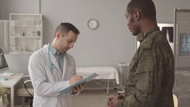 Medium slowmo of African American army officer in uniform and Caucasian male doctor in lab coat with clipboard having conversation during appointment at modern doctors office