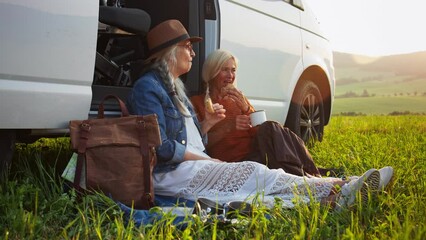 Senior women friends sitting by car boot outdoors at sunset, caravan trip holiday. - Powered by Adobe