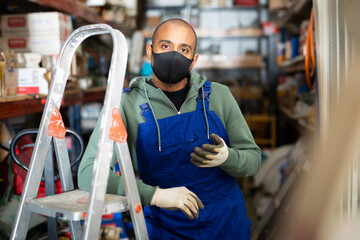 Portrait of hispanic worker of building materials store in protective mask to prevent viral infections posing near stepladder in wareroom