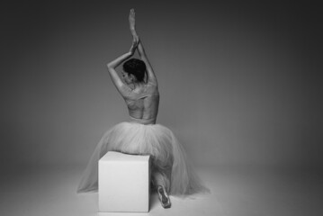 classic black and white photo, a young pretty, fragile, beautiful ballerina sitting on a white square, rolling up her arms in a long dress with a tulle on a uniform background. Ballet, dance, dancer