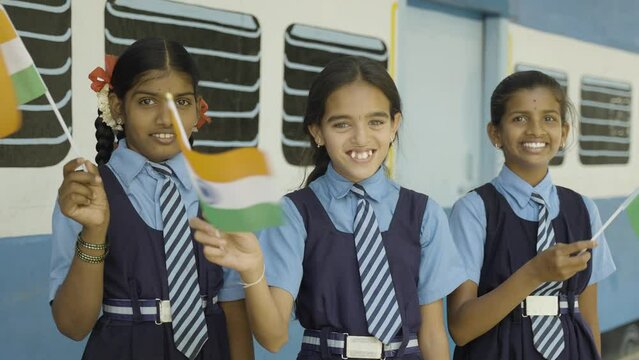 Smiling girl kis in unifrom waving Indian falg by looking at camera at school corridor - concept of Patriotism, republic or independence day celebration