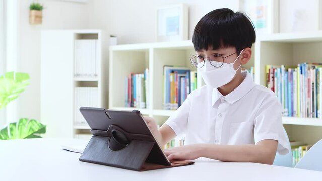 Boy wearing a mask, studying via internet online with laptop and tablet and taking network video lessons at home