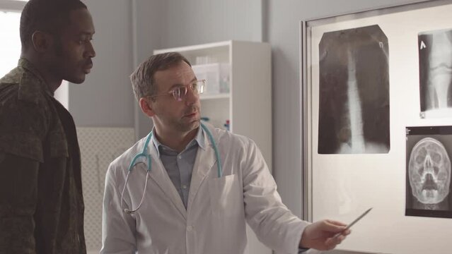Medium slowmo of radiologist and African American army officer standing by projector with patients x rays having appointment at modern clinic. Doctor analyzing results and telling diagnosis