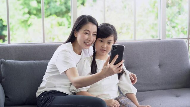 Portrait young Asian mother and daughter using smartphone for broadcasting livestream or having video call, waving hand, saying hello, goodbye via video call or FaceTime feeling happy to cellphone.