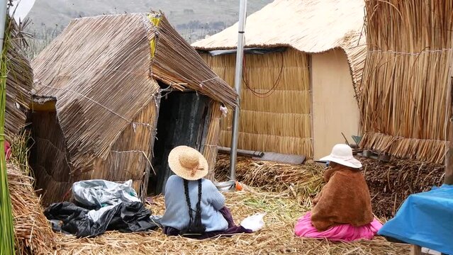 Two Indigenous Women In Hats Sitting Near A Traditional Reed House In Peru