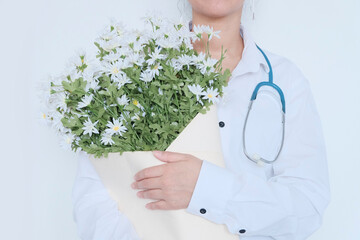 The doctor is a woman of Asian appearance, with black hair, in white medical clothes, holding a bouquet of chamomile flowers in her hands. The concept of congratulations on the day of the medic, nurse