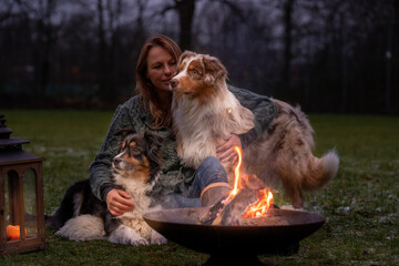 Young woman is sitting outside in the woods with her two Australian Shepherd dogs. Snow on the grass, at night by the campfire. She embraces the dogs lovingly
