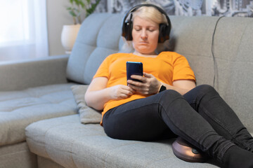 A girl in headphones and with a phone in her hands, sitting on the couch, makes a foot massage with a massage pillow.