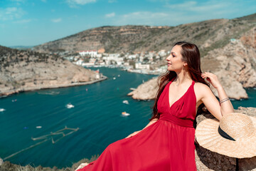 A woman, a side view in a red flying dress fluttering in the wind, a girl in a fluttering dress on the background of the sea. A straw hat hangs at the back of the neck. The concept of a vacation at