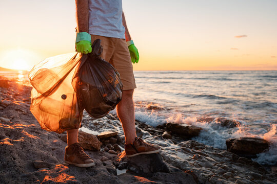 A male volunteer holding two polyethilene bag with garbage. Close up. In the background the sea and the sunset. Copy space. The concept of environmental protection and Earth day
