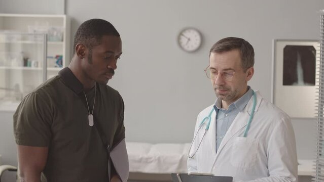 Tilt up medium slowmo portrait of injured African American army officer with broken arm in sling and Caucasian male medical worker talking then looking at camera standing at modern doctors office
