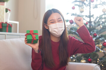Asian woman with face mask receiving Christmas presents and unpacking box, sitting on sofa in cozy living room. Holiday season in Covid pandemic.