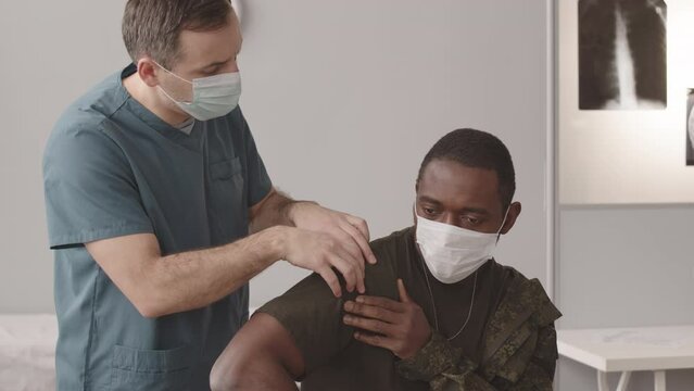 Medium slowmo of Caucasian male army doctor examining injured African American soldier with shoulder dislocation, patients xrays on projector in background