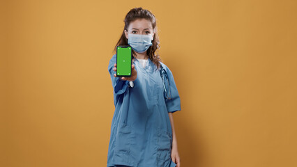 Doctor with stethoscope holding smartphone and texting showing green screen mockup at camera. Medic in hospital uniform wearing surgical mask using touchscreen device presenting copy space.