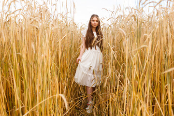 Woman in white dress standing in field with spikelets in summer and looking at camera 