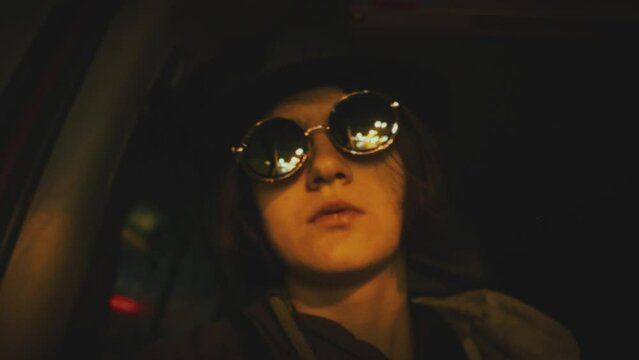 Girl wearing a hat and mirror sunglasses reflecting colorful city lights, night driving in a car. Slow motion. Vintage stylized video with step print and slight film grain effect.