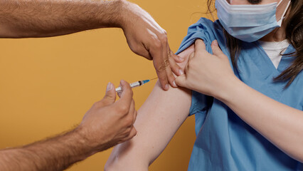 Closeup of medical doctor wearing surgical mask lifting sleeve and recieving covid or flu vaccine in studio. Detail view of medic wearing virus protection getting immunization shot from syringe.
