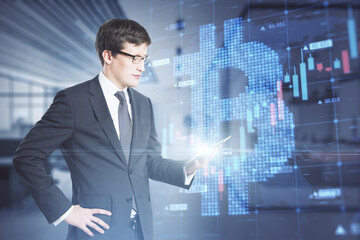 Attractive young caucasian businessman holding tablet with creative glowing bitcoin grid forex chart hologram on blurry office interior background. Cryptocurrency and growth concept. Double exposure.