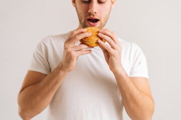 Close-up shot of hunger young man holding eating delicious burger on white isolated background. Studio shot of happy handsome male bitting tasty unhealthy meal.