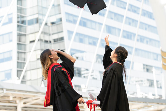 Happy two Asian young beautiful graduate female students with master and bachelor degree throwing cap to the sky after graduation while holding diploma in hand. Blur background of University building