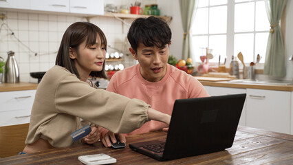 smiling asian husband and wife pointing at computer while discussing what to buy on the internet at home. they pay online with a credit card