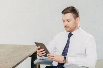Horizontal shot of handsome young successful male fiinancier uses modern tablet computer, wears white shirt with tie, has break after work, sits at wooden table in his cabinet, reads information
