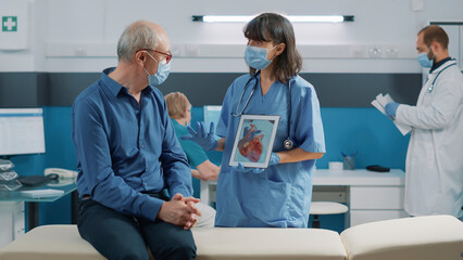 Nurse and elder patient looking at cardiology illustration on digital tablet during coronavirus pandemic. Woman and man analyzing cardiovascular system and heart condition on screen.