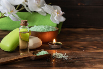 Body oil in a clear bottle, fragrant soap and bath salt on a dark table. Green bath towel and orchid sprig in the background