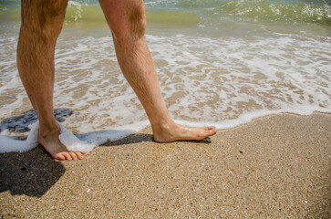 Men's feet stand on the sandy shore and in white foam from the waves