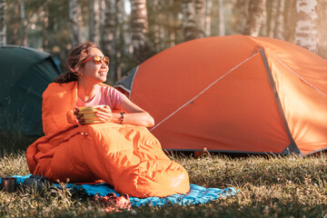 Woman in a sleeping bag eating breakfast against the backdrop of many tents in a camping site at...