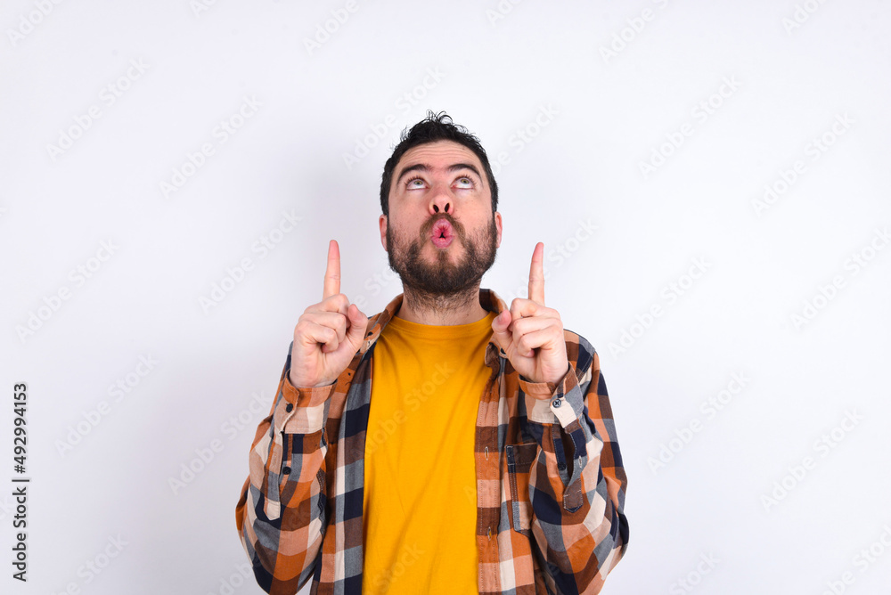 Poster young caucasian man wearing plaid shirt over white background being amazed and surprised looking and pointing up with fingers showing something strange.