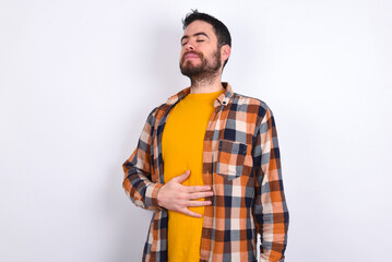 Satisfied smiling young caucasian man wearing plaid shirt over white, keeps hands on belly, being...